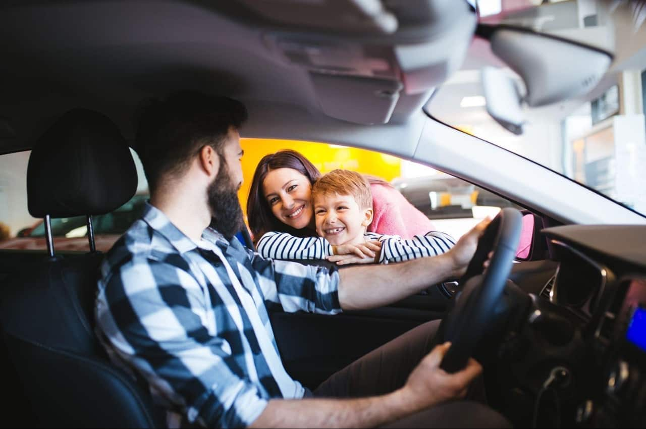 Mãe e filho sorridentes, apoiados na janela de motorista de veículo, olham para o pai testando um carro. 