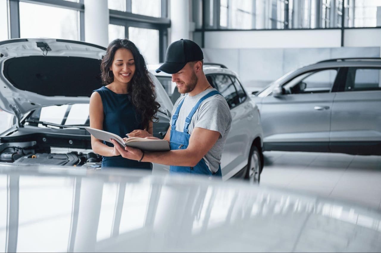 Homem mostrando vistoria veicular para uma mulher. Eles estão em uma concessionária de carros, onde um deles está com o capô aberto.