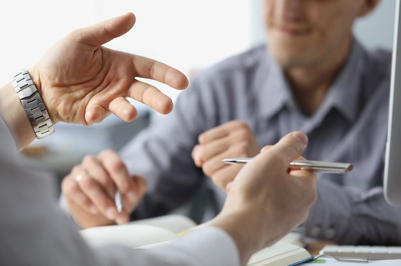 Dois homens conversando em escritório. Seus rostos não aparecem na imagem e eles estão sentados em uma mesa.