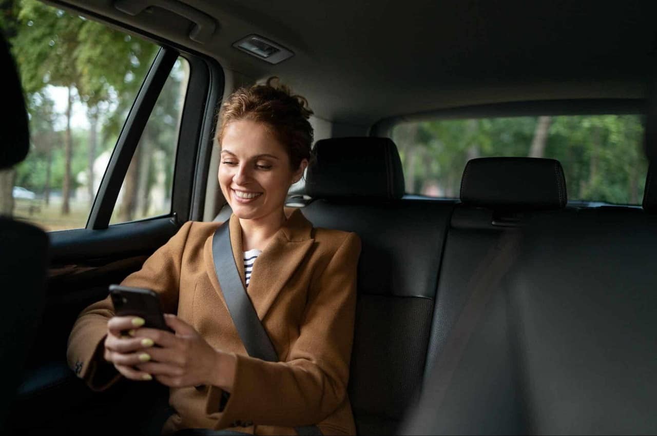 Mulher sorridente no banco de trás do passageiros usando app de transporte no celular.