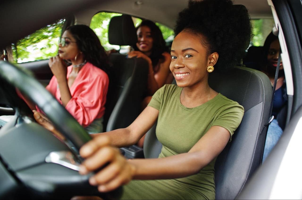 Mulher negra sorridente de blusa verde, dirigindo um veículo e com duas passageiras mulheres no carro.