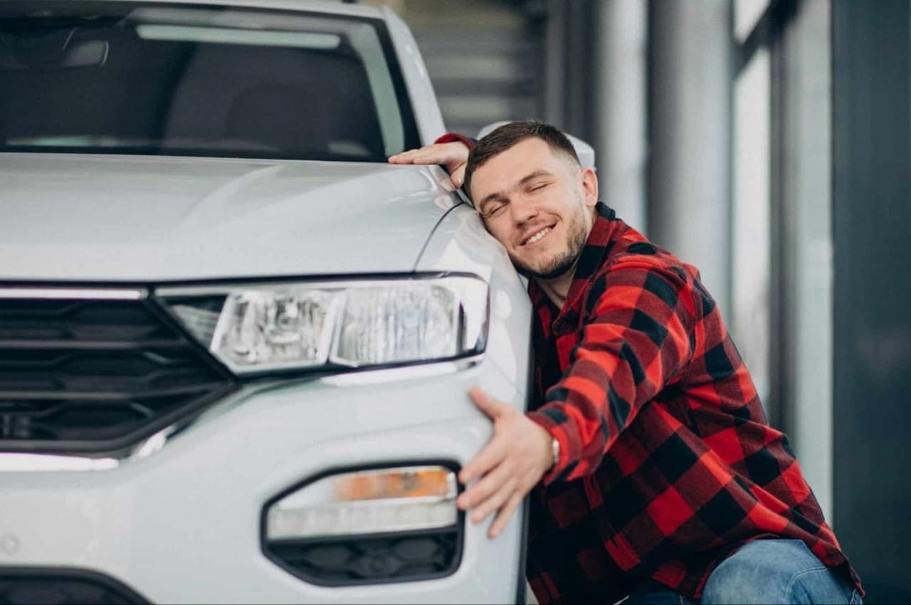 Homem com expressão sorridente, vestindo blusa xadrez vermelha e abraçando um carro prata na concessionária.