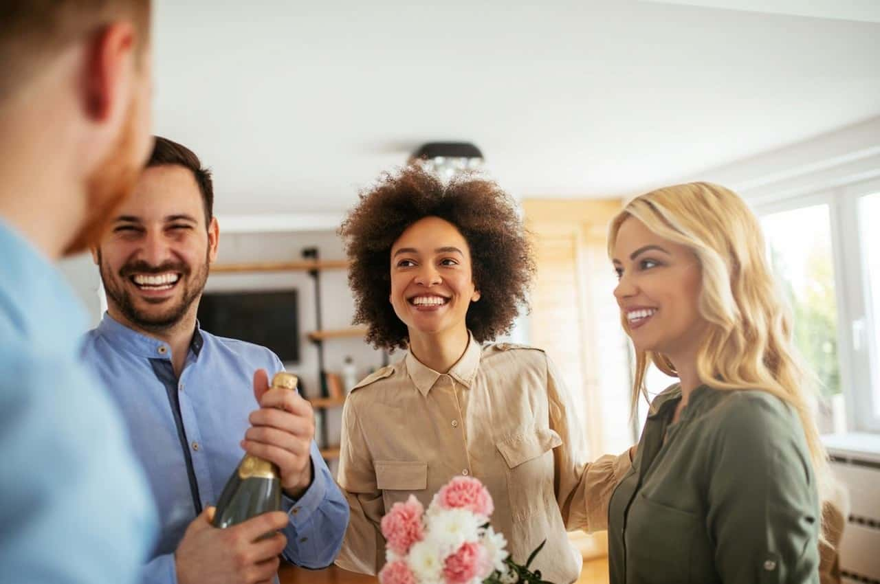 Grupos de amigos sorridentes carregando flores e garrafa de champanhe fechada para comemorar compra de apartamento.