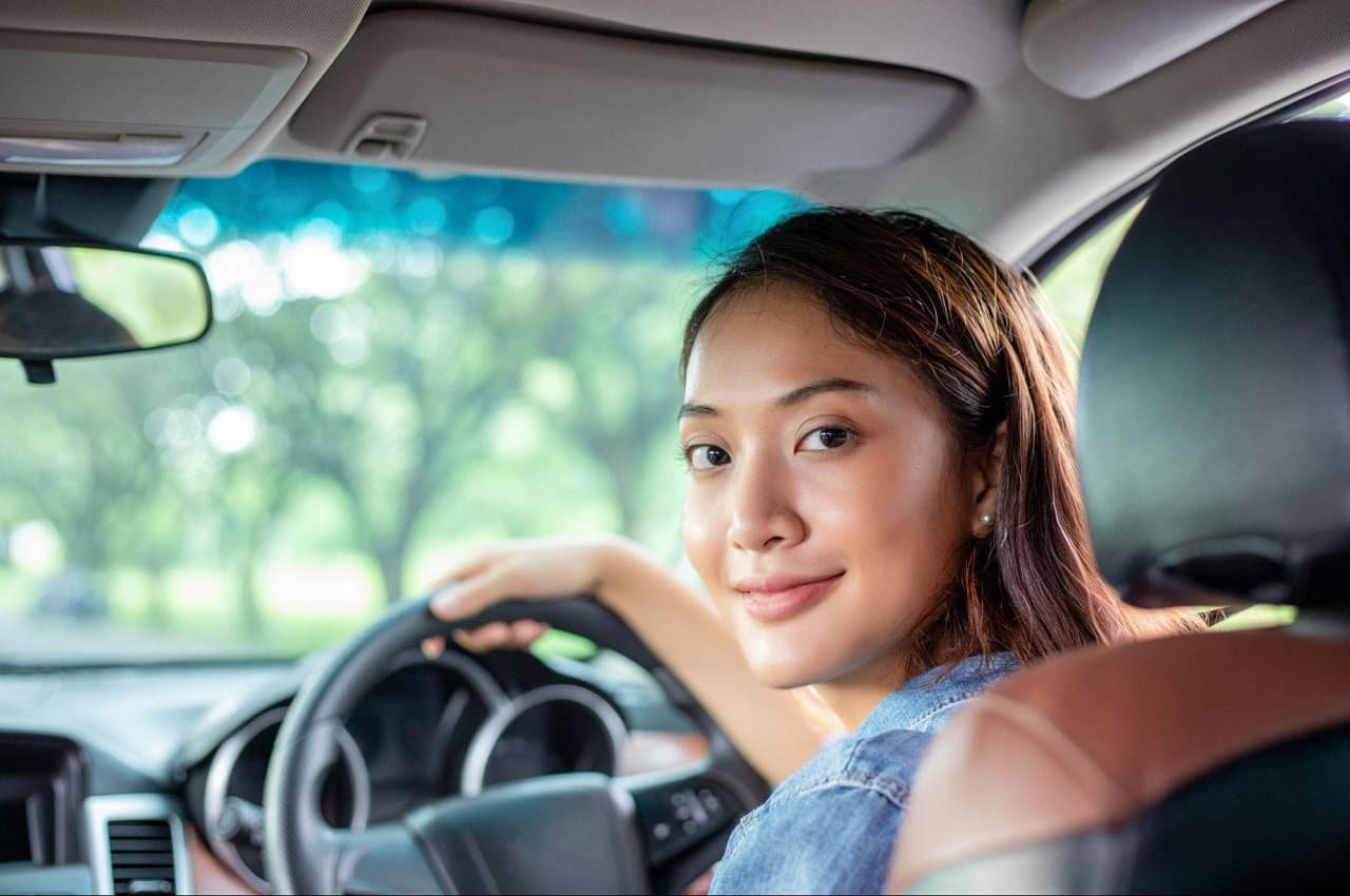 Mulher sorridente, no banco de motorista e segurando volante do veículo. Ela olha para trás.