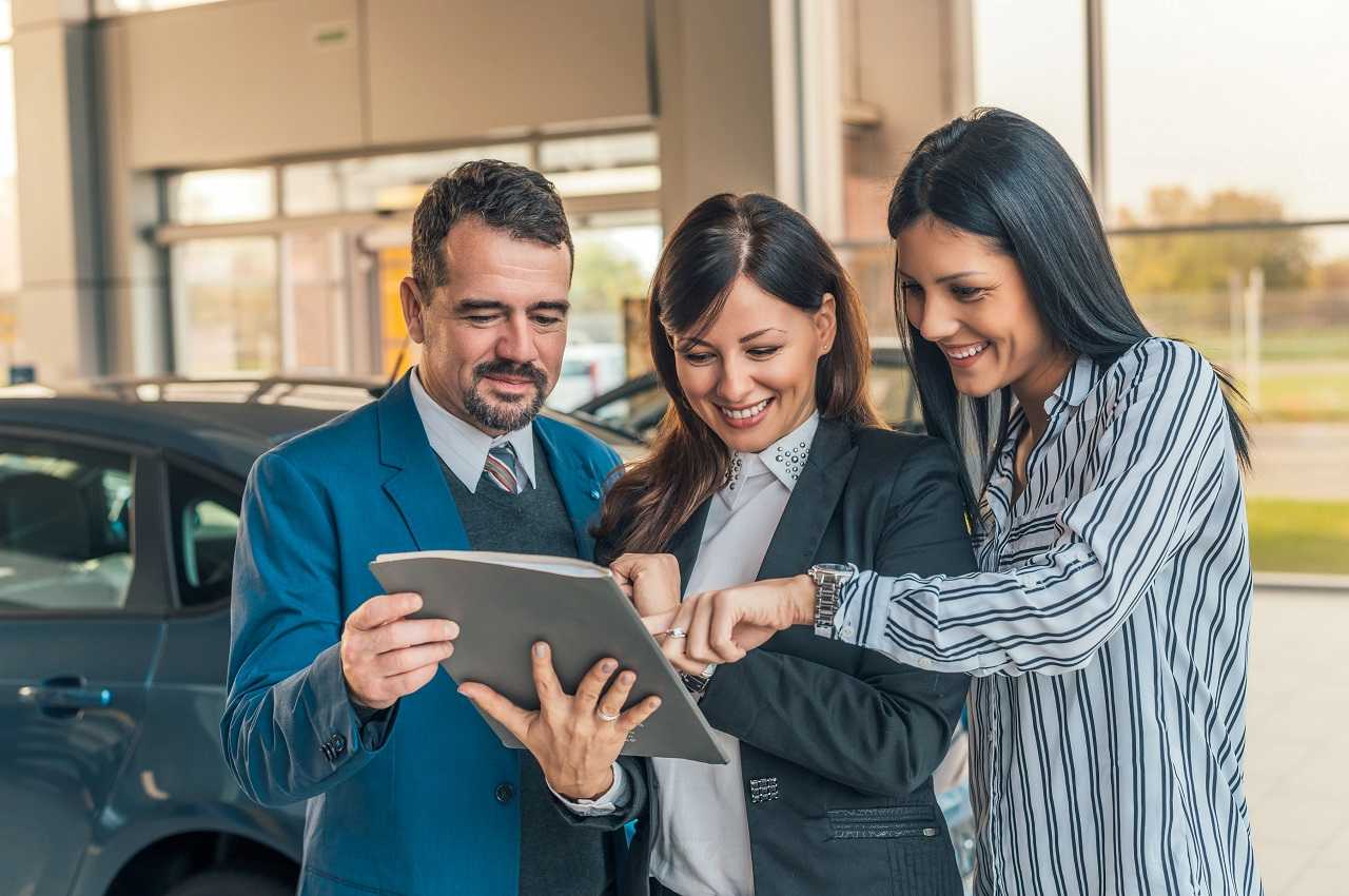 Duas pessoas (homem e mulher) comprando carro próprio com vendedora de concessionária que segura uma prancheta.