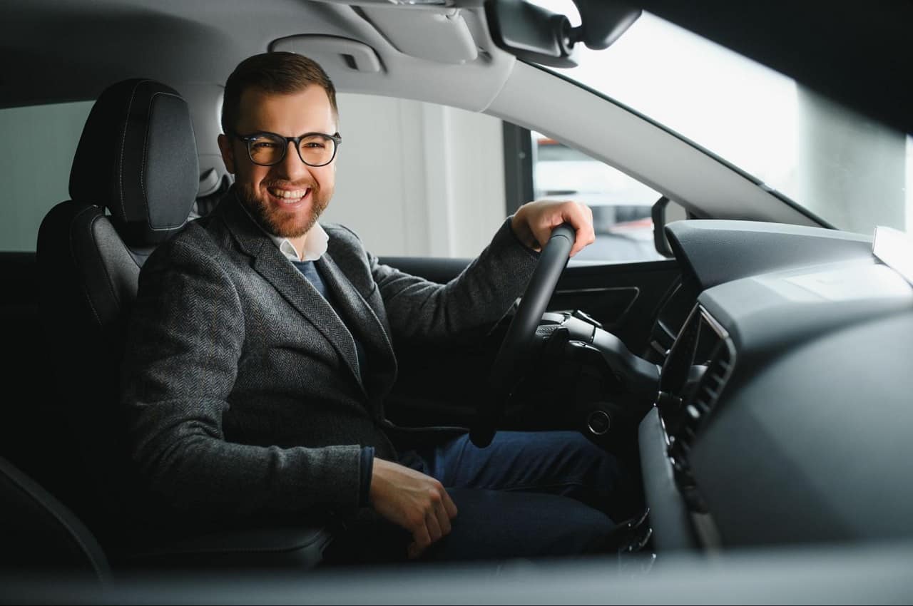 Homem sorridente vestindo terno de lã, óculos de grau e dirigindo um veículo moderno.