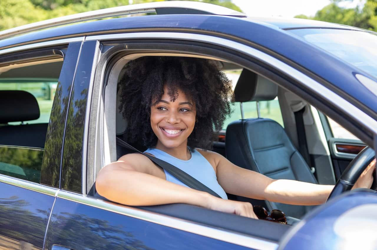 Jovem de cabelo crespo sorrindo, enquanto segura o volante de um carro financiado sem entrada.