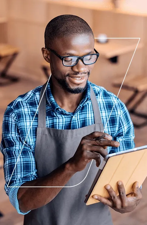 Homem sorrindo, vestindo um avental de cozinha dentro de um restaurante, segurando um tablet para anotação de pedidos