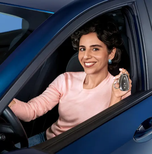 A imagem mostra uma mulher de pele branca sorrindo e olhando pela janela do seu carro, no banco do motorista. Com a mão direita ela segura o volante e com a mão esquerda está segurando e mostrando a chave do carro. Ela tem o cabelo escuro, curto e encaracolado, está usando um brinco discreto de formas geométricas em tons azuis e uma blusa na cor rosa salmão.