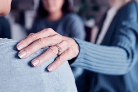 A imagem mostra uma mão com um anel, repousando suavemente no ombro de alguém que está vestindo uma blusa azul-clara.