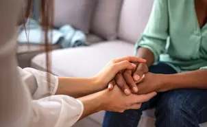 Duas pessoas de mãos dadas em sofá cinza-claro. Uma de blusa branca, pele clara; outra, de camisa azul, pele negra.