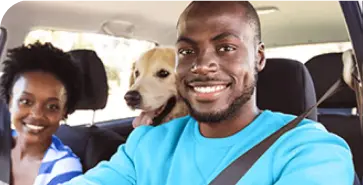 Na imagem um homem negro veste blusa azul e ocupa o assento do motorista. Ao lado vemos uma mulher negra e um cão.