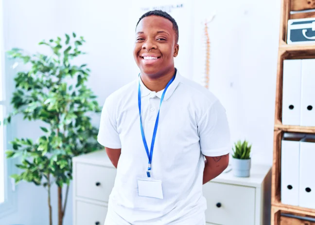 A imagem mostra um homem em um escritório claro. Ele está sorrindo, veste uma camiseta branca e usa um crachá.