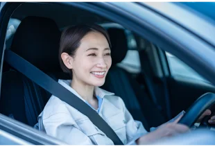 A imagem mostra um mulher sentada dentro de um carro, segurando o volante, enquanto sorri.