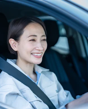 A imagem mostra um mulher sentada dentro de um carro, segurando o volante, enquanto sorri.