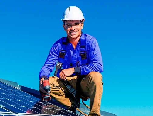 A imagem mostra um homem branco, de capacete branco, blusa de manga longa azul e calça bege. Ele está em cima de um painel solar, segurando um objeto de trabalho na mão direita e representa um integrador. O céu está azul e ensolarado.