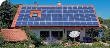 Casa com painéis solares, cercada por árvores e flores, e à frente, uma pequena casinha com telhado marrom.