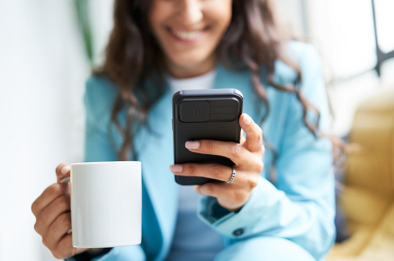 A imagem mostra uma mulher, segurando uma caneca de porcelana e usando o seu celular. 