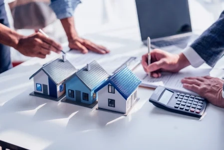 Na imagem há três modelos de casas em miniatura em uma mesa, pessoas anotando em papéis, laptop e calculadora ao lado.