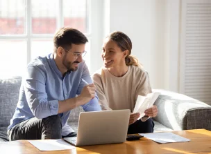 Duas pessoas sentadas olham para papéis e um laptop. A mulher é branca de cabelos escuros e o homem tem pele clara e barba.