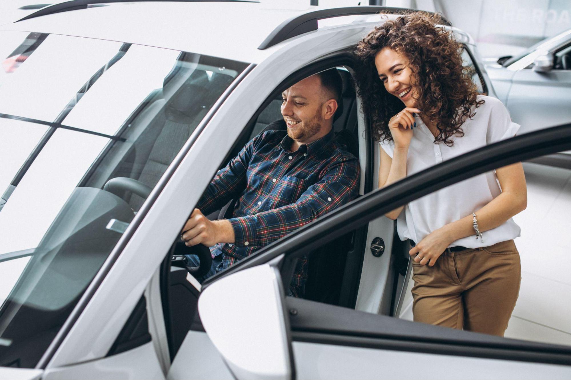 A imagem mostra um homem dentro de um carro com a porta do motorista aberta. Ao lado dele, uma mulher, em pé e encostada no carro. Ambos estão sorrindo.