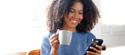 Mulher com cabelos cacheados e camisa azul, sorrindo, segurando uma caneca branca com uma mão e um celular com a outra.