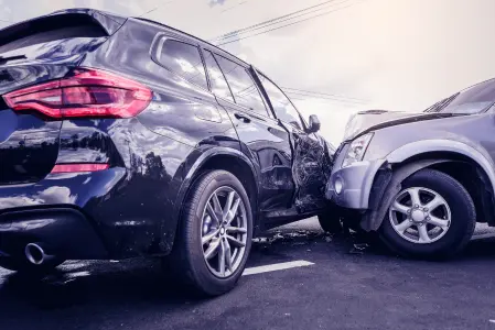 A imagem mostra dois carros, sendo um escuro e um claro e ambos amassados após uma colisão em estacionamento.