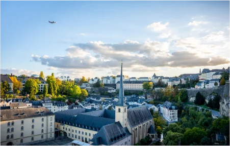 Vista superior da cidade de Luxemburgo, com uma igreja cercada de edifícios e arvores, um céu azul claro com nuvens e um avião sobrevoando a cidade.