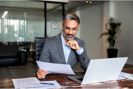 Homem com cabelos e barba grisalhos, de terno cinza e óculos de grau, olhando para um papel em uma mesa de escritório, com um notebook aberto â frente.