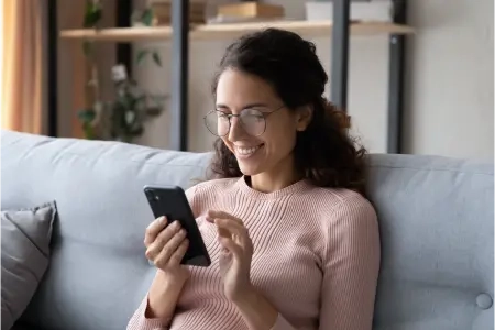 Na imagem há uma mulher sentada em um sofá, ela está mexendo no celular sorrindo.