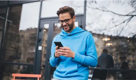 Na imagem há um homem de óculos de grau, ele está em um local aberto, trajado com moletom azul, sorrindo para um celular.