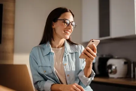 Na imagem há uma mulher de cabelos longos e escuros segurando um celular sorrindo.