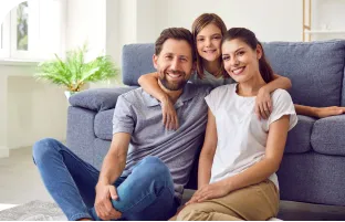 Homem e mulher sentados no chão em uma sala de estar, enquanto uma menina sentada em um sofá azul abraça os dois.